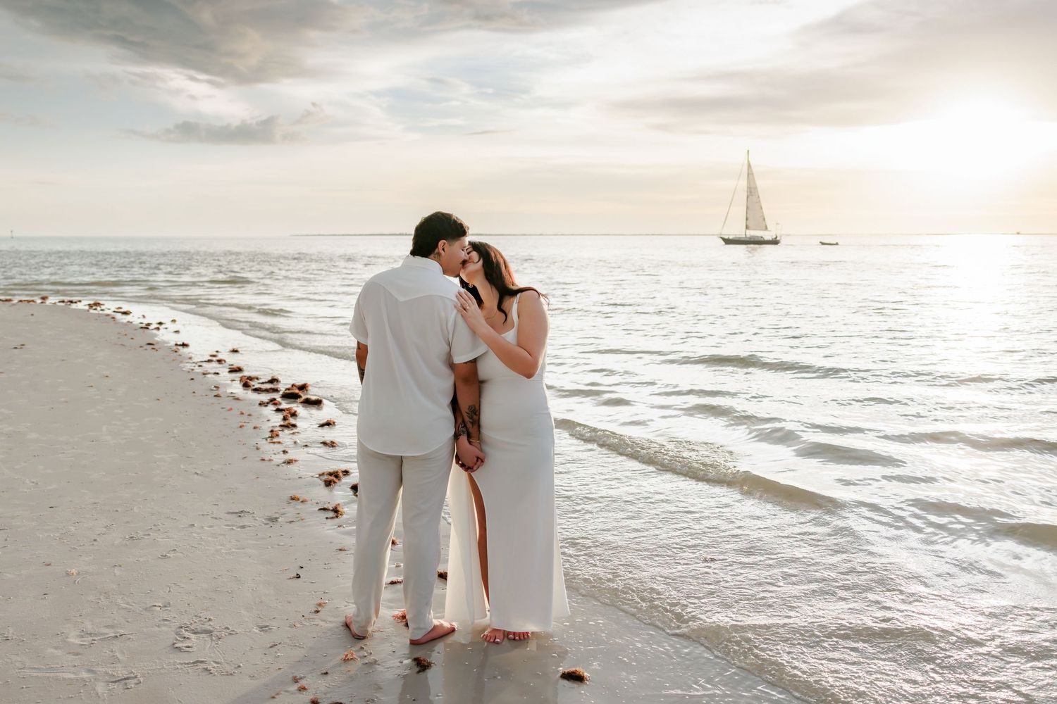 Couple in white embracing on a sandy beach at sunset with a sailboat on calm ocean water in the background.