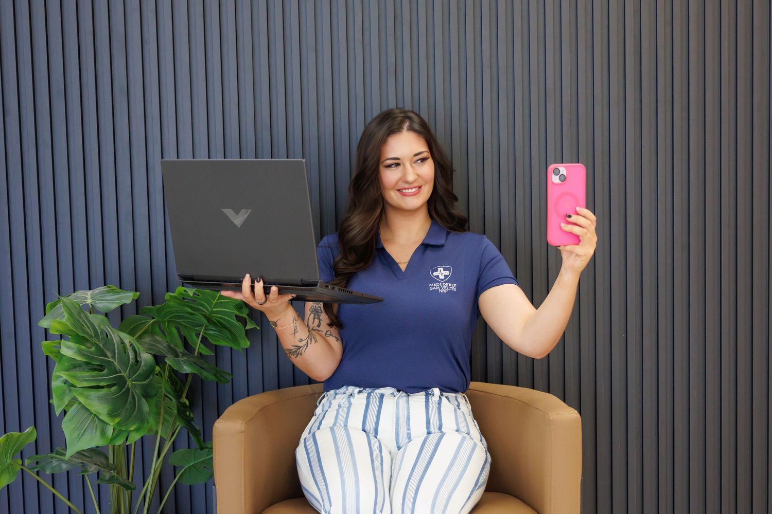 Woman in navy polo holds laptop and pink smartphone while sitting in modern office chair near green plant.