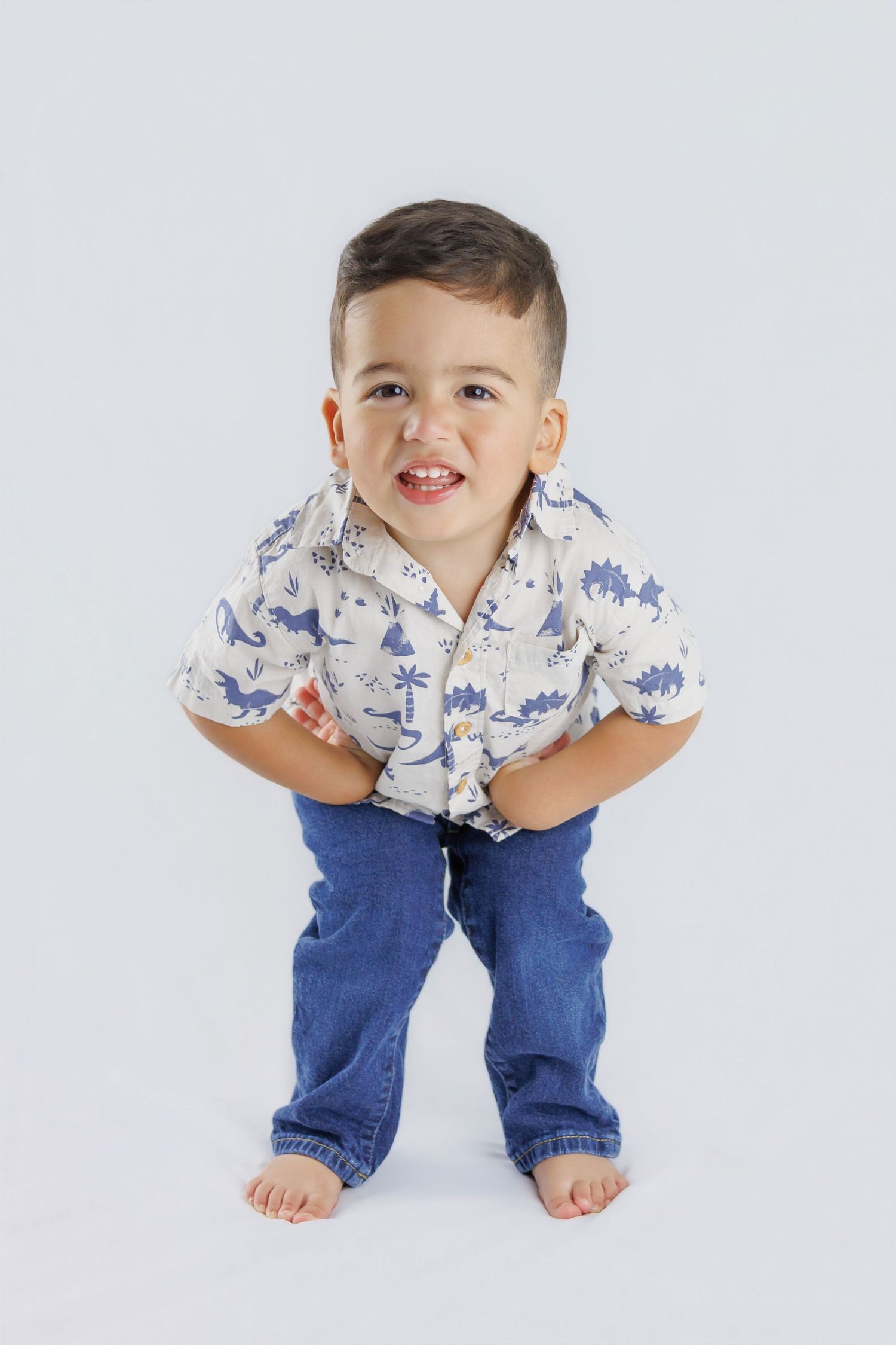 Playful toddler boy in a dinosaur shirt and jeans leaning forward and smiling against a white background.