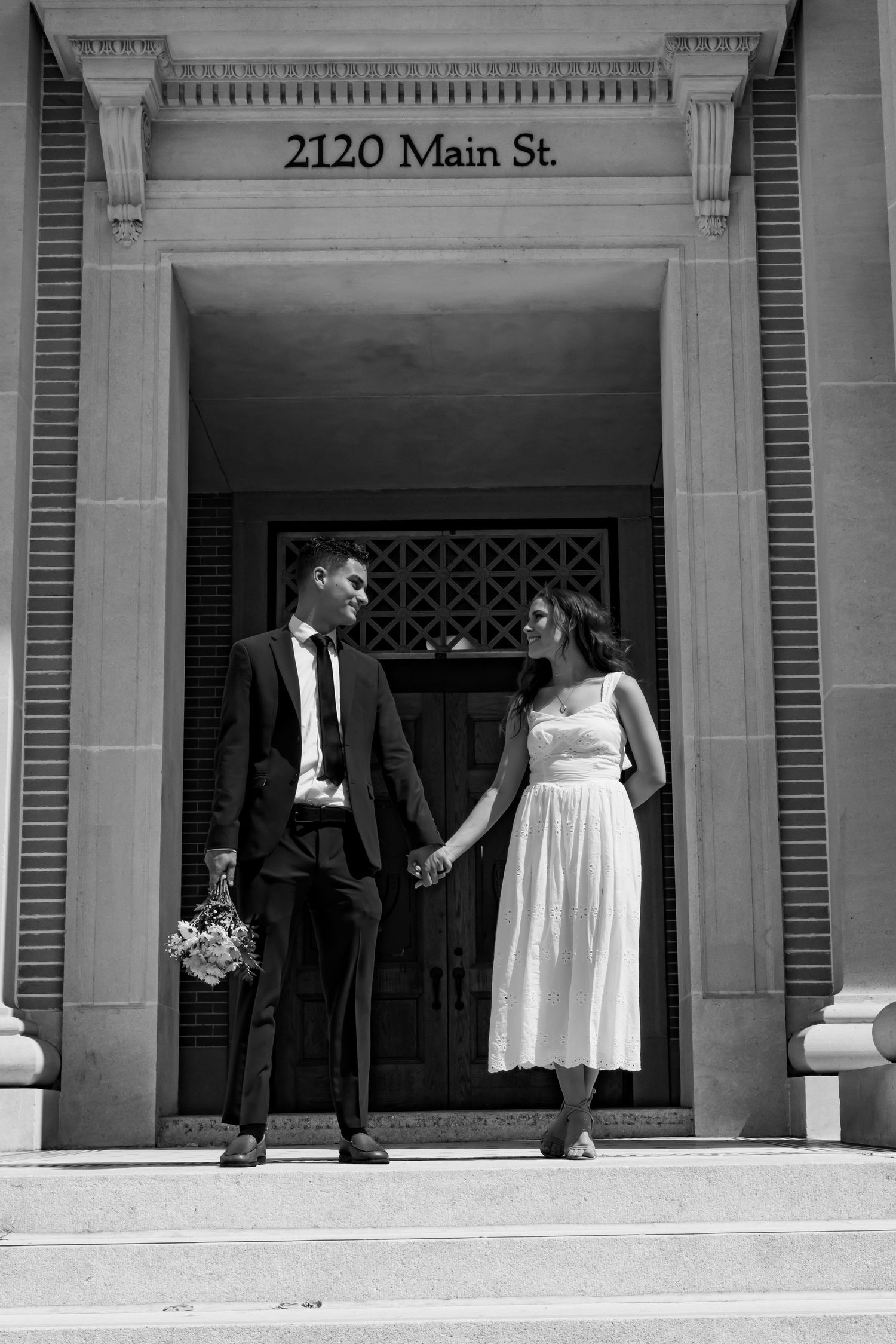 Black and white photo of a couple holding hands on steps at 2120 Main St., bride in white dress and groom in suit.