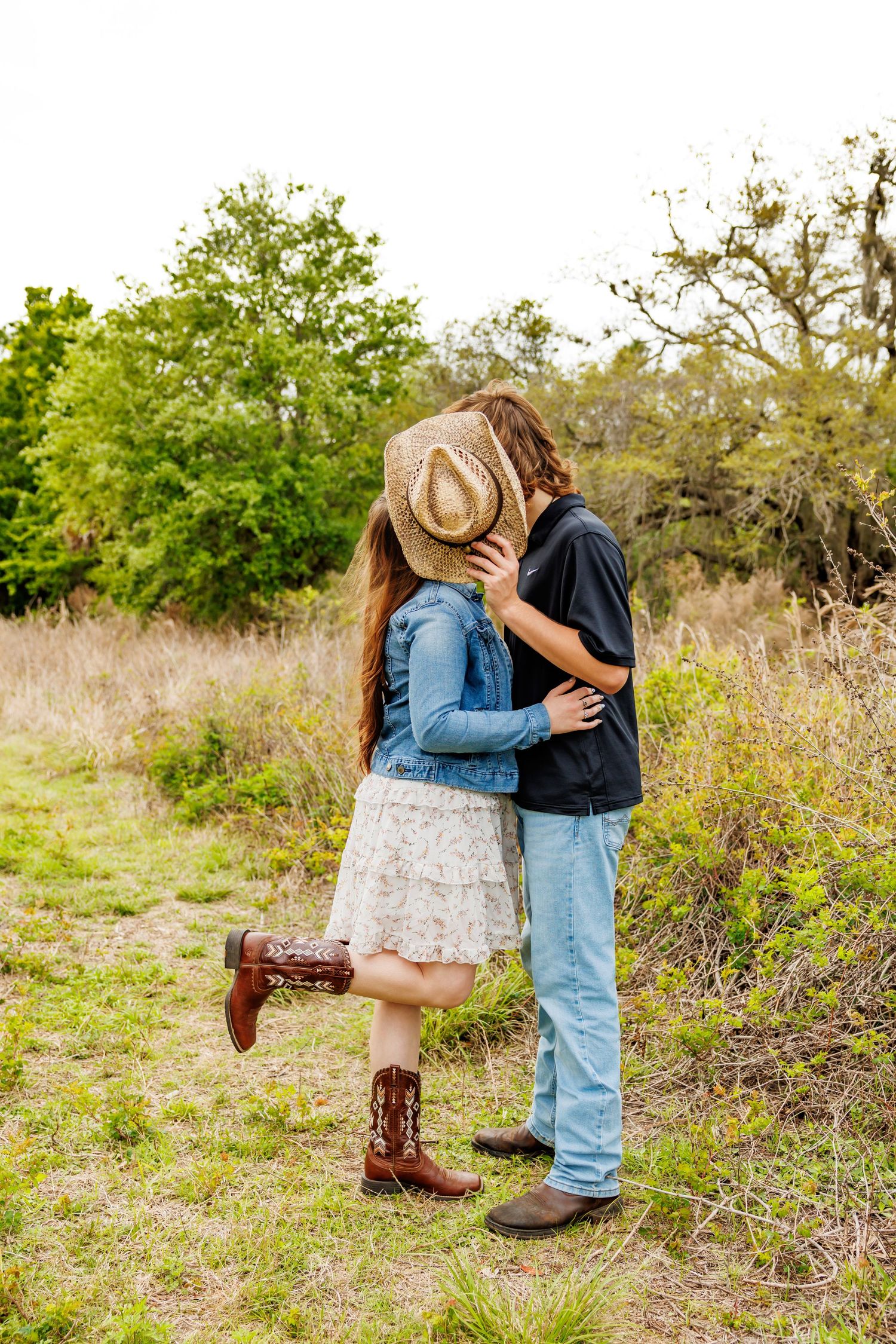Couple kissing in a field, woman in cowboy boots and denim jacket holds a straw hat over their faces.