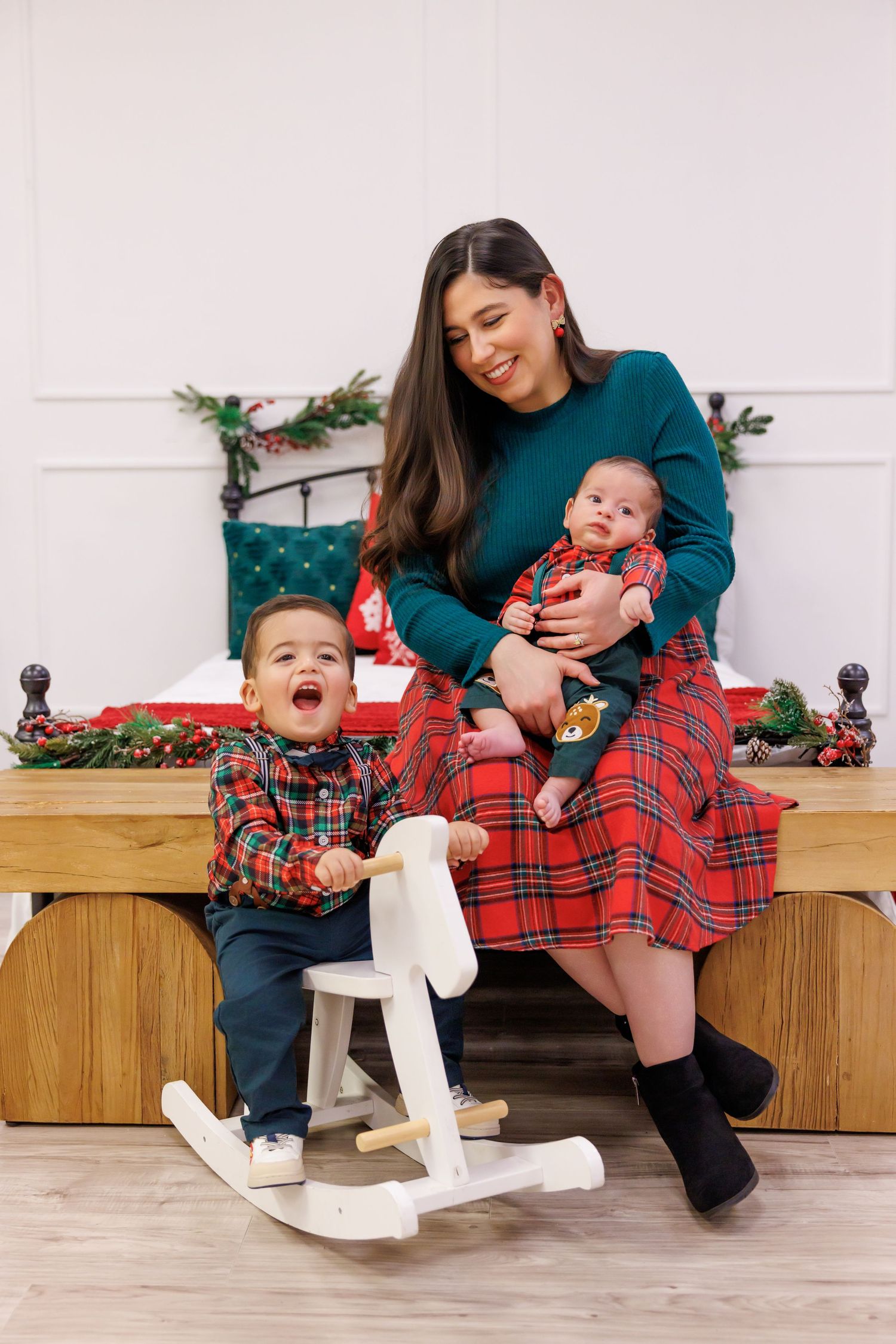 A smiling mother in a plaid skirt holds her newborn while her toddler son plays with a rocking horse in Christmas decor.