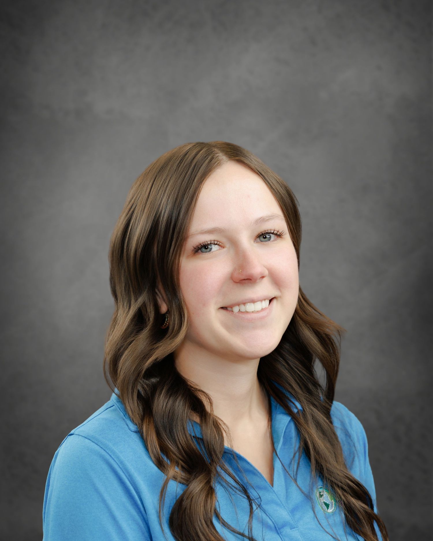 Smiling woman with long wavy brown hair wearing a blue polo shirt in a professional headshot against a gray background.