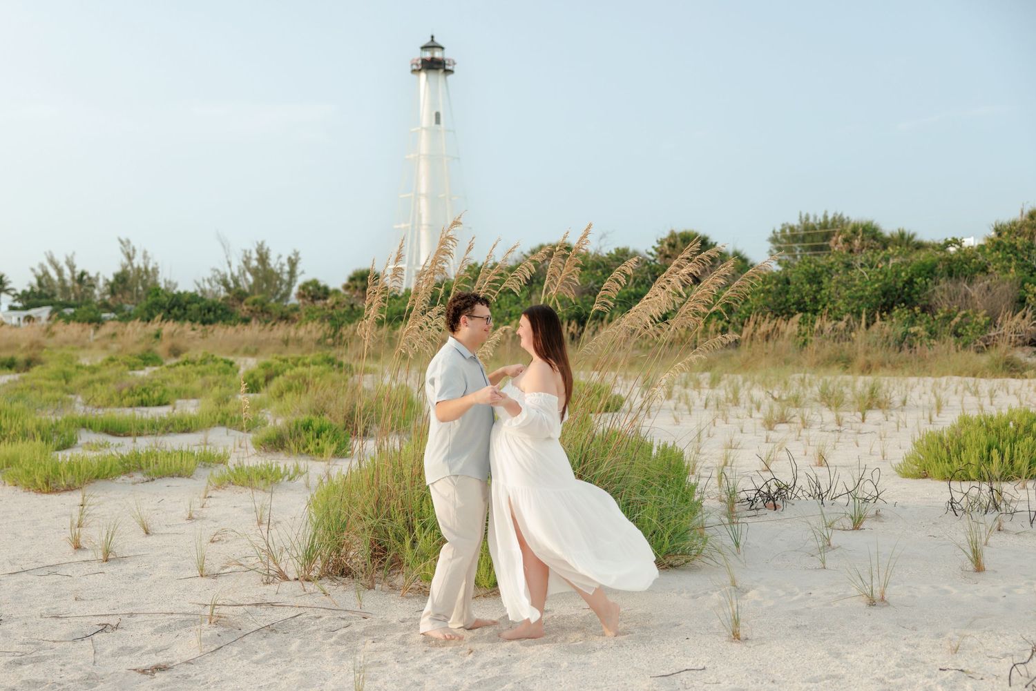 Couple in white embracing and dancing on a sandy beach with sea oats and a tall white lighthouse in the background.