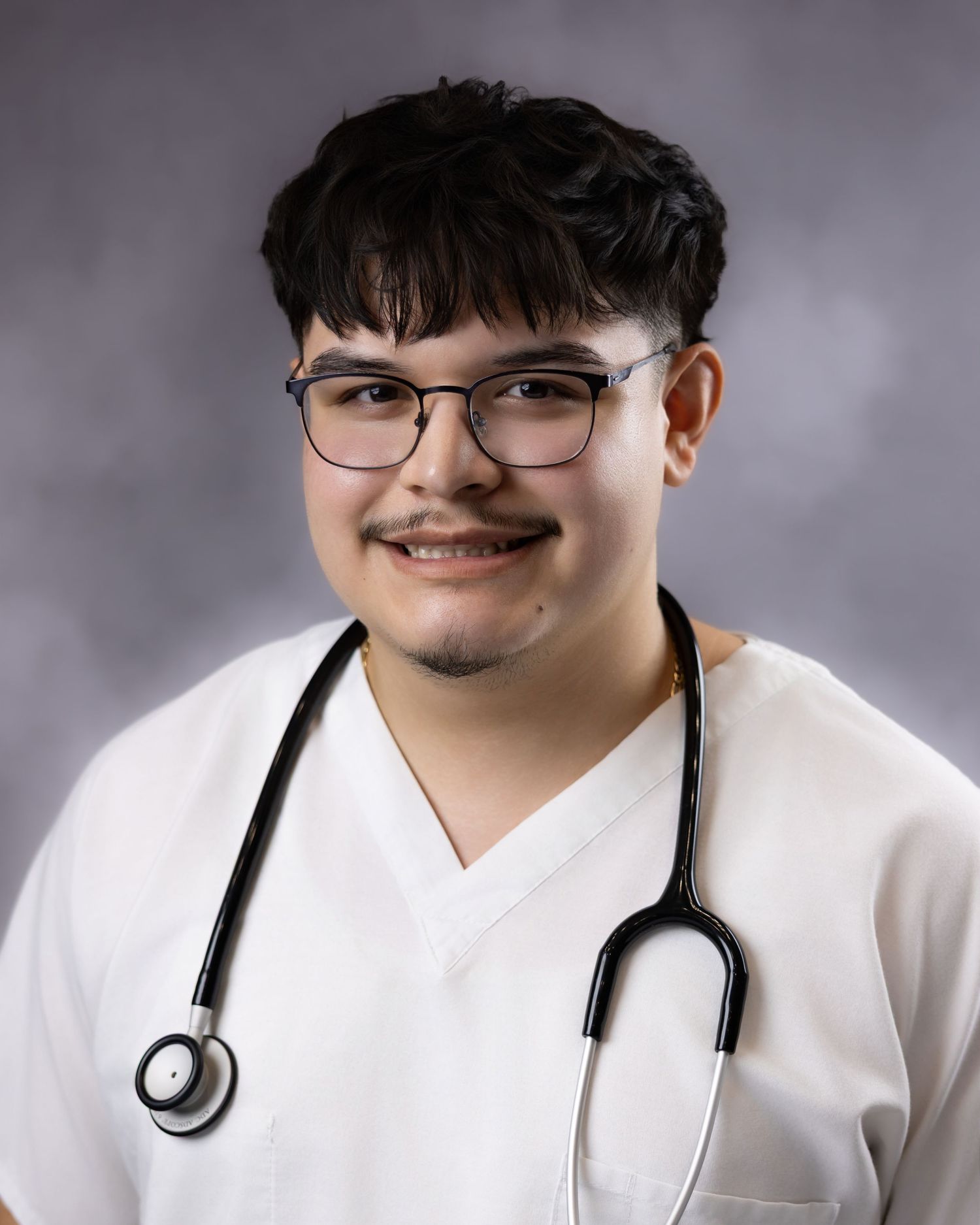 Young male healthcare professional in white scrubs with stethoscope and glasses smiling in a studio portrait.