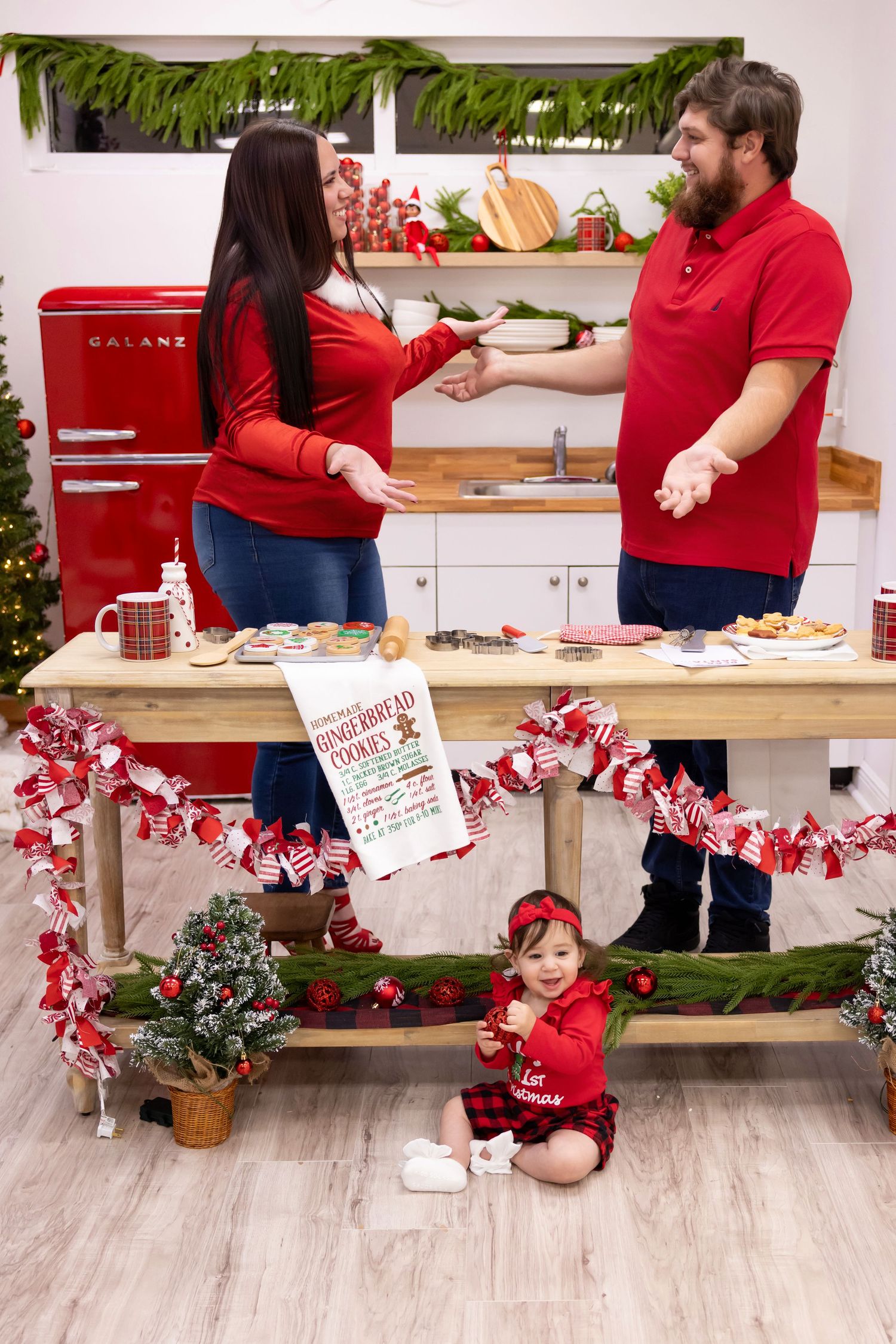 Family in matching red outfits baking Christmas cookies in a festive kitchen with a toddler sitting on the floor.