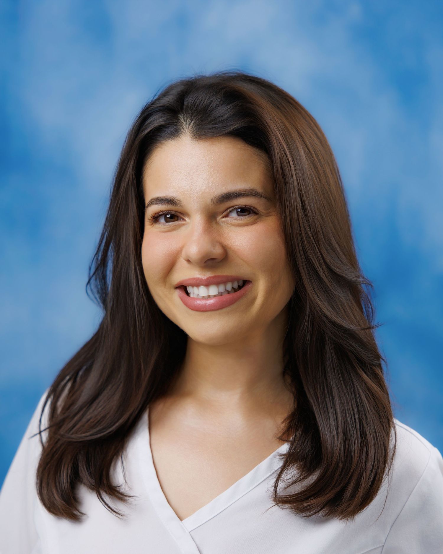 Professional headshot of smiling brunette woman in white top against blue background.