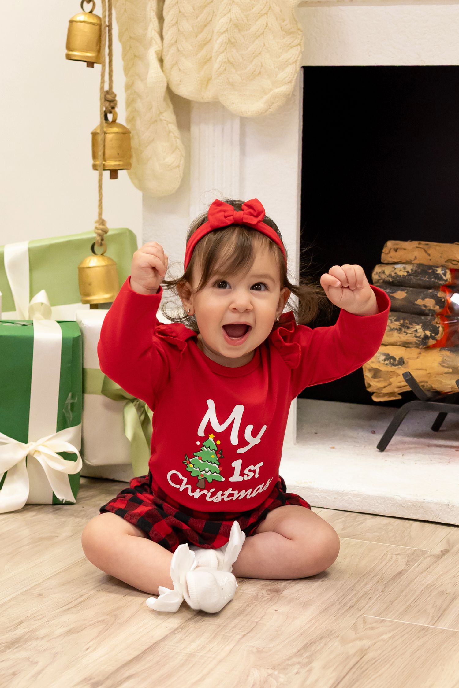 Excited baby girl in red My 1st Christmas outfit sits before a fireplace with gifts and holiday decorations.