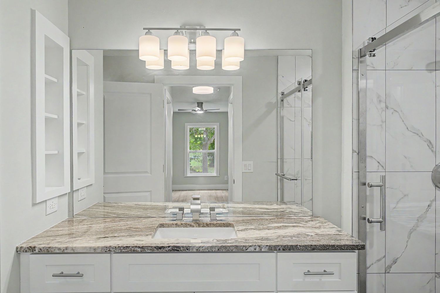 Modern bathroom with a marble-top vanity, white cabinetry, a three-light fixture, large mirror and a glass-enclosed marble shower.