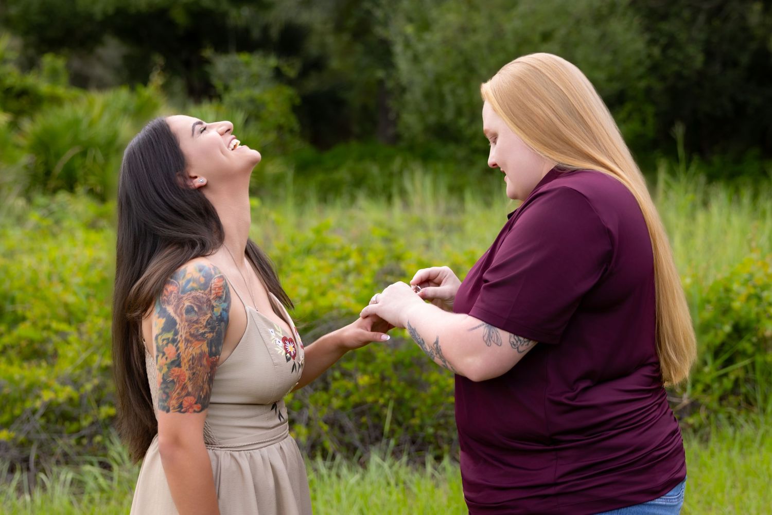 Two women sharing a joyful proposal moment outdoors, one with tattoos laughing as the other places a ring on her finger.