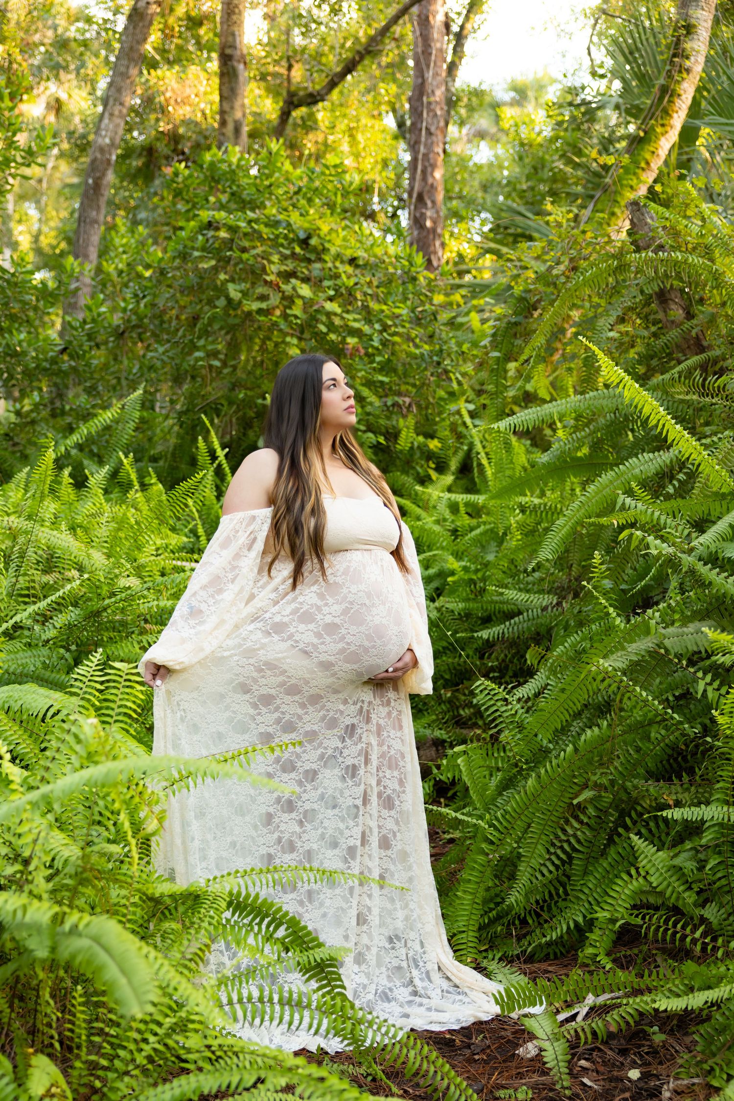 Pregnant woman in white lace gown cradling baby bump, surrounded by lush green ferns in a forest setting.
