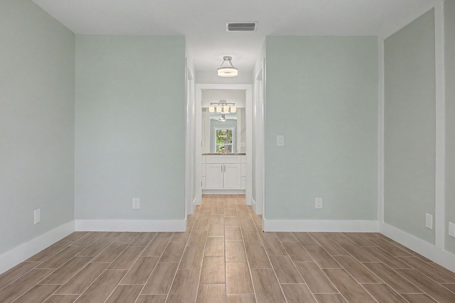 Empty room with light sage green walls and wood-look tile flooring leading to a hallway and a bathroom with a vanity.