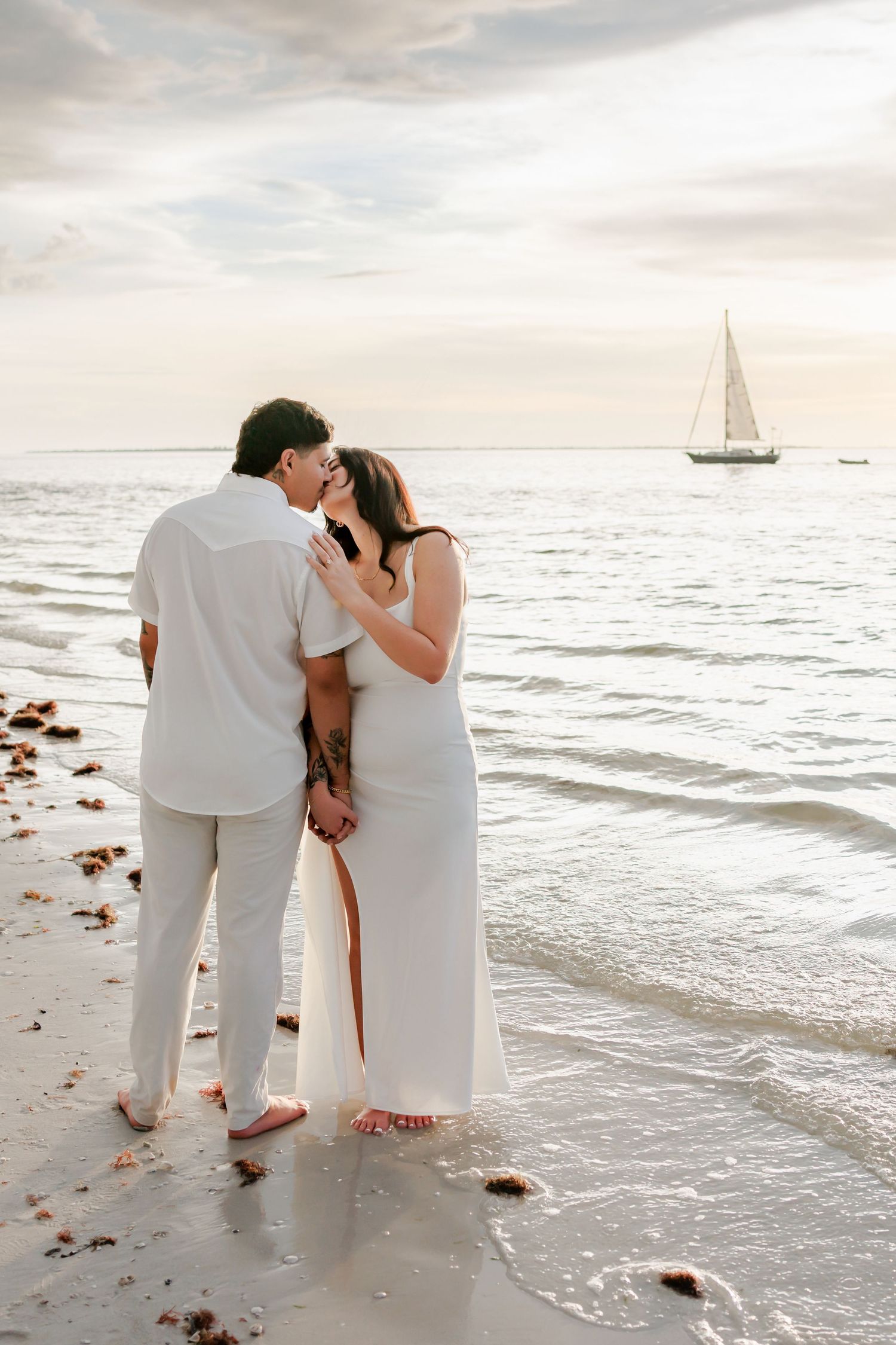 Barefoot couple in white embracing and kissing on a sandy beach at sunset, with a sailboat on the calm water behind them.
