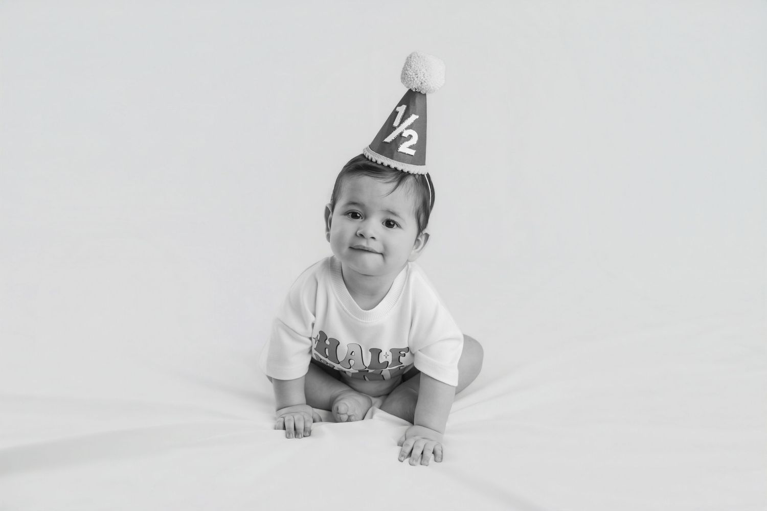 Black and white photo of a baby in a half birthday hat sitting on a white backdrop.