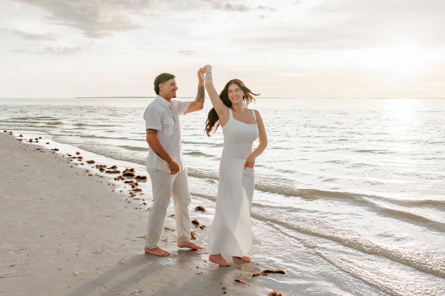 Couple in white outfits dancing hand in hand on a sandy beach at sunset, with gentle waves rolling in behind them.