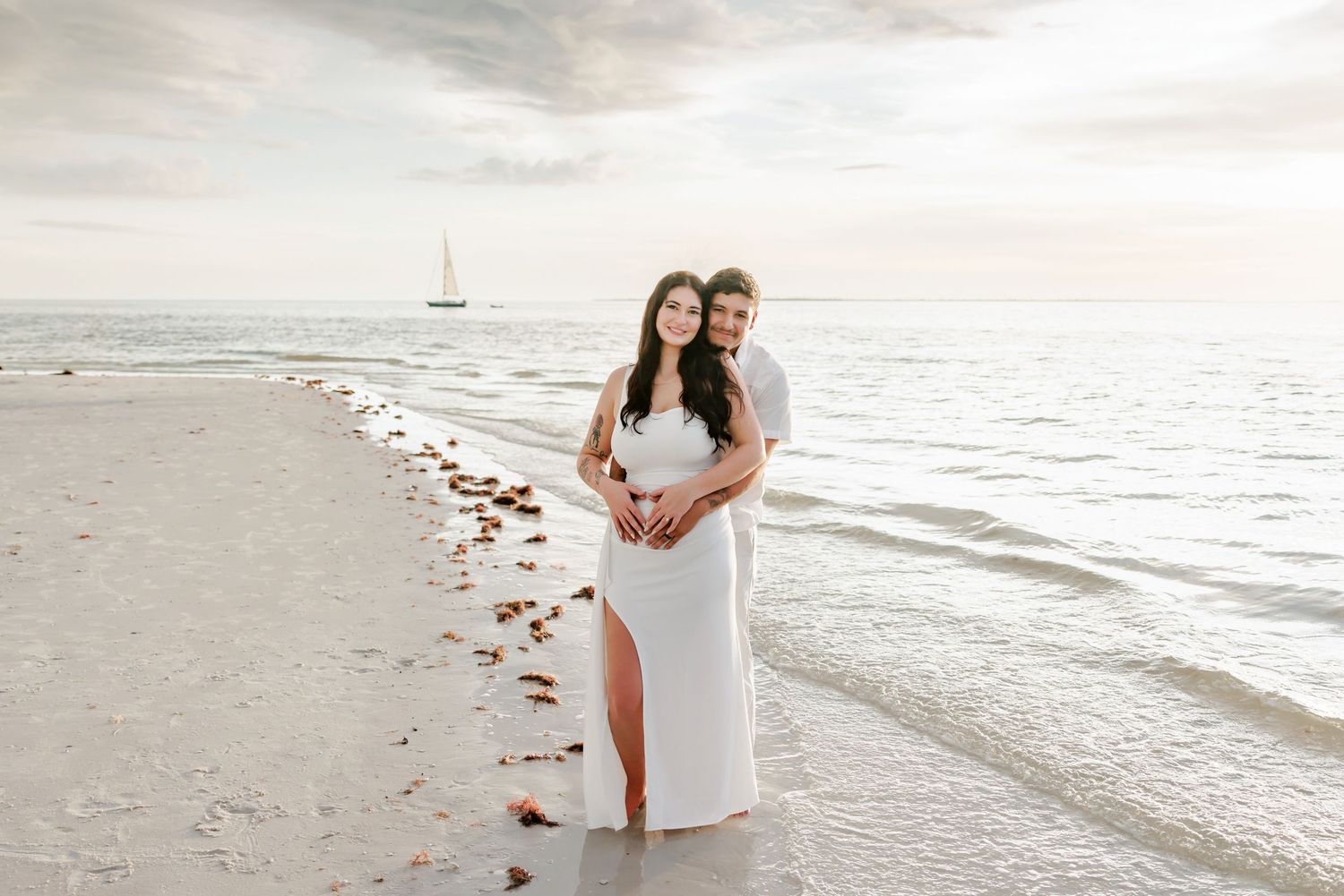 Couple embracing on a sandy beach at sunset, the woman in a white dress with a leg slit, with a sailboat on the calm ocean behind.