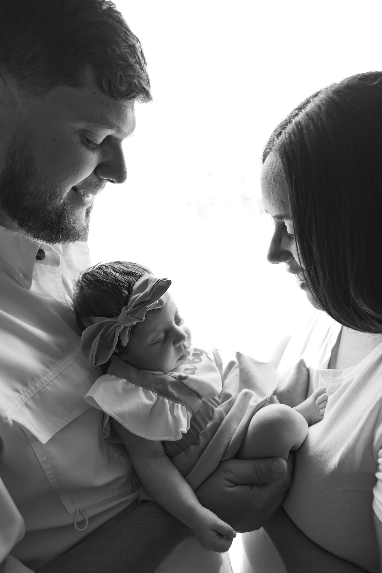 Black and white photo of parents lovingly gazing at their newborn baby girl wearing a floral headband.