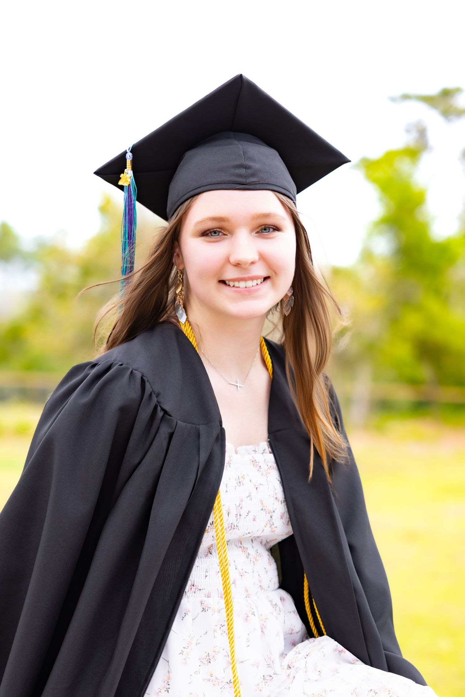 Smiling young woman in black graduation cap and gown poses outdoors with green trees in background.