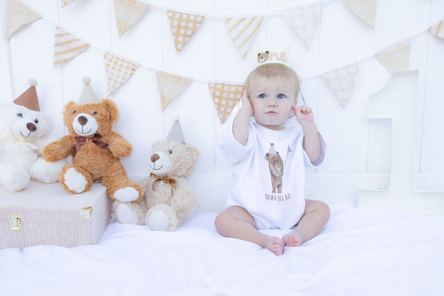 Baby in birthday onesie sits with teddy bears and bunting decorations for a first birthday photo shoot.