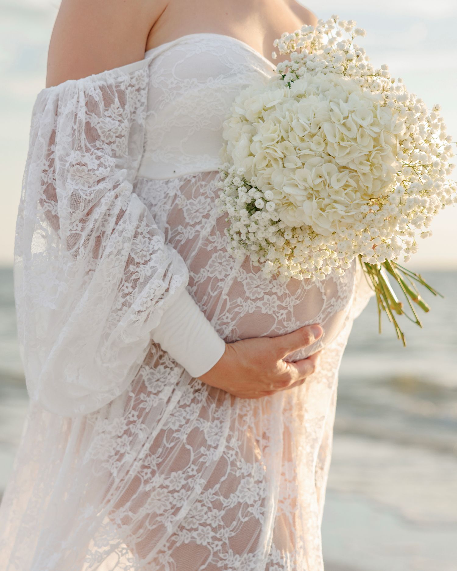 Woman in a white lace off-the-shoulder maternity gown holding a white hydrangea and baby's breath bouquet against her belly on a beach.