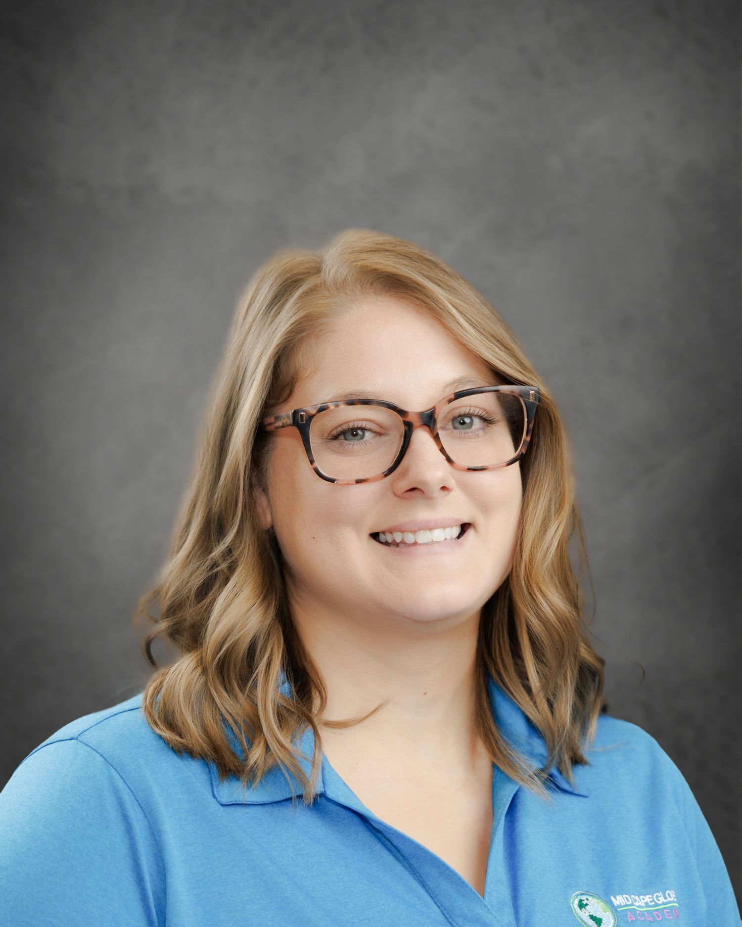Smiling woman with shoulder-length wavy blonde hair and tortoiseshell glasses wearing a light blue polo shirt in a professional headshot.