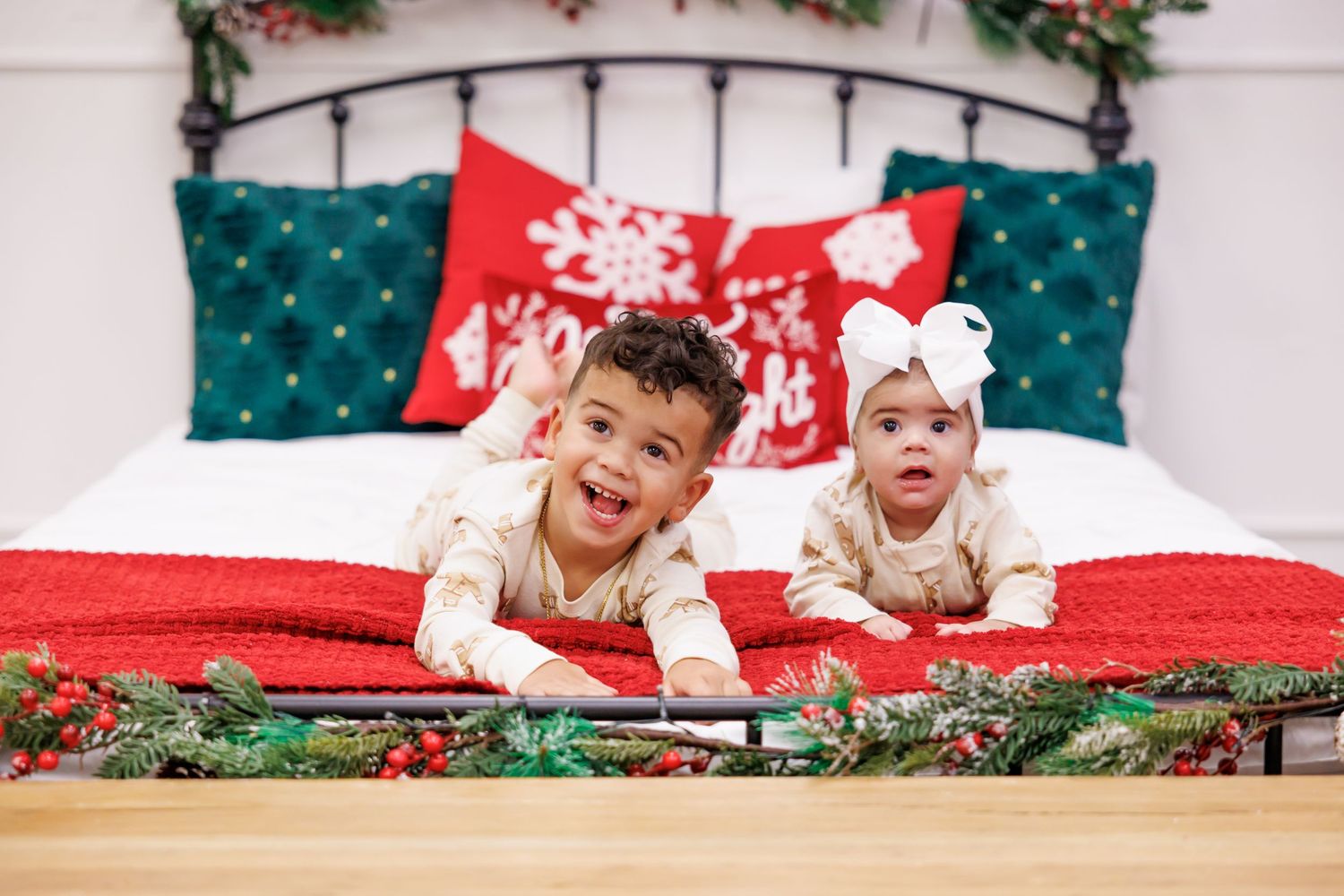 Two young children in holiday pajamas lying on a red Christmas-decorated bed with festive pillows and garland.