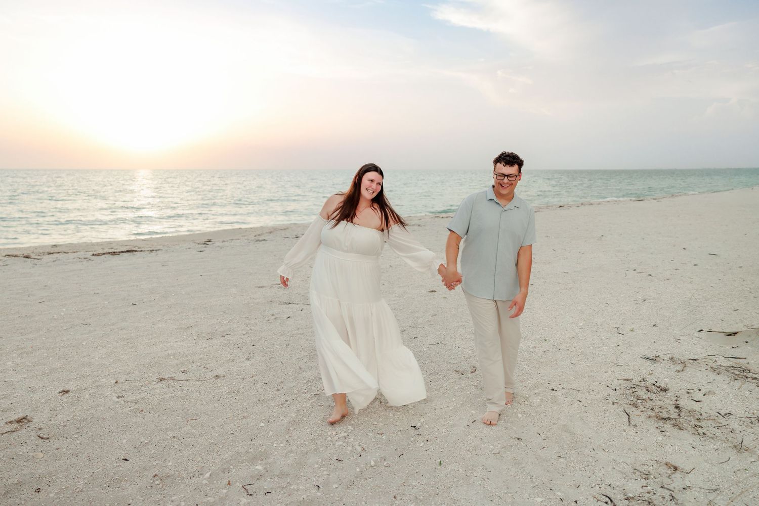 Smiling couple holding hands and walking barefoot along a sandy beach at sunset, the woman in a flowing white dress beside the calm ocean.