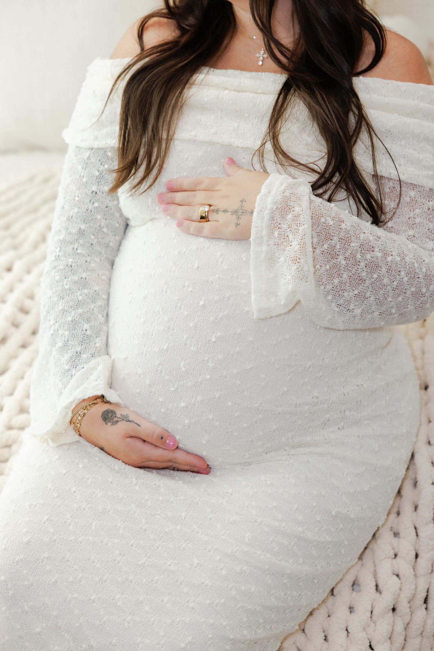 Pregnant woman in a white off-the-shoulder lace maternity gown resting her hands on her baby bump on a cozy chunky knit blanket.
