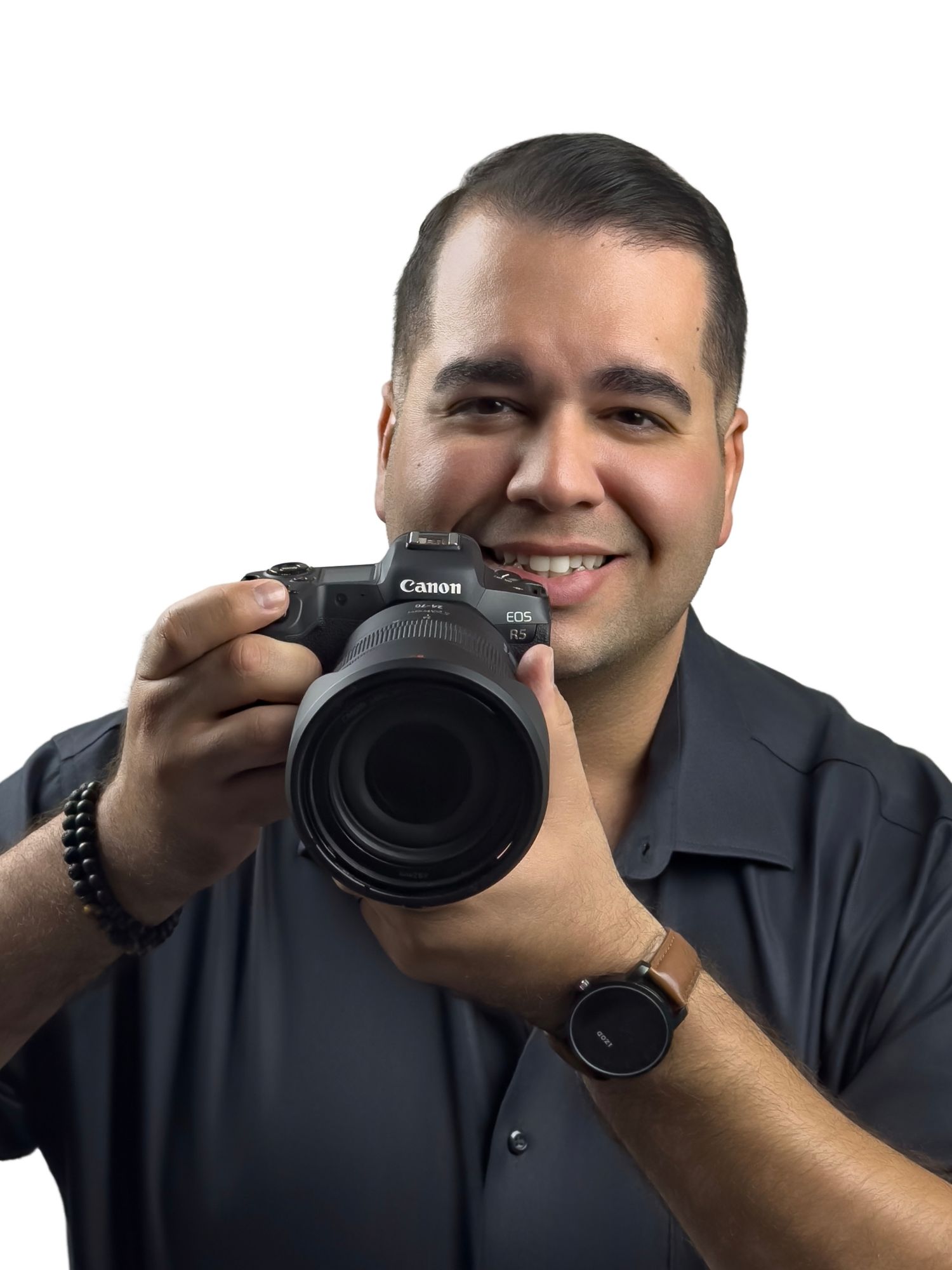 Smiling man holding a Canon EOS camera toward the viewer, wearing a gray shirt and watch, on white background.
