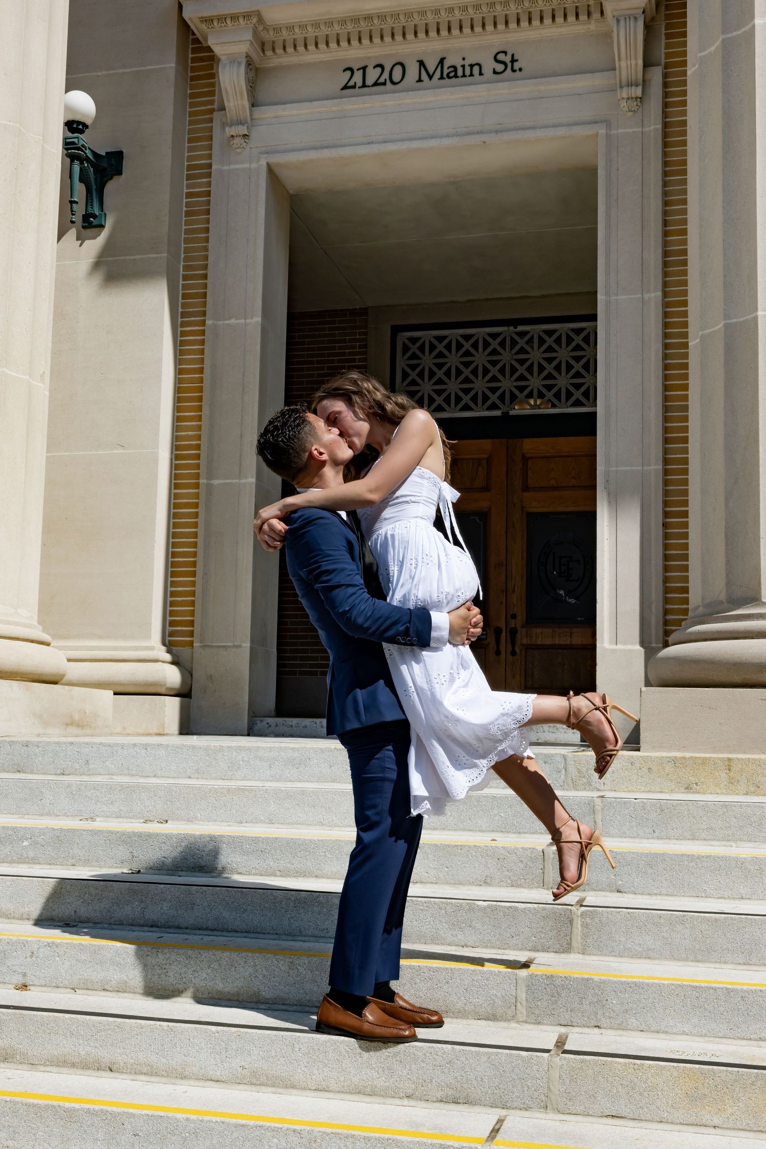 Couple sharing a romantic kiss on the steps of 2120 Main St., groom lifting bride in white dress and blue suit.