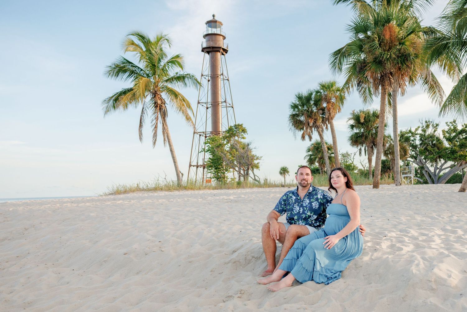Couple sitting together on a sandy beach with palm trees and a historic lighthouse in the background during a sunny coastal photo session.