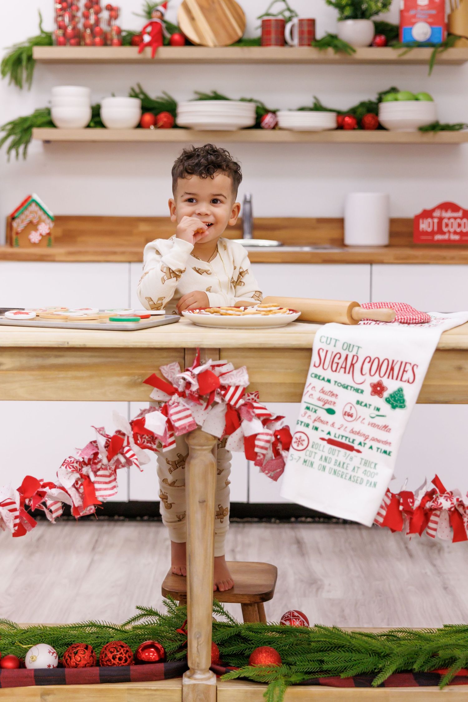 Toddler boy eating cookie at decorated Christmas kitchen table with sugar cookie recipe towel.