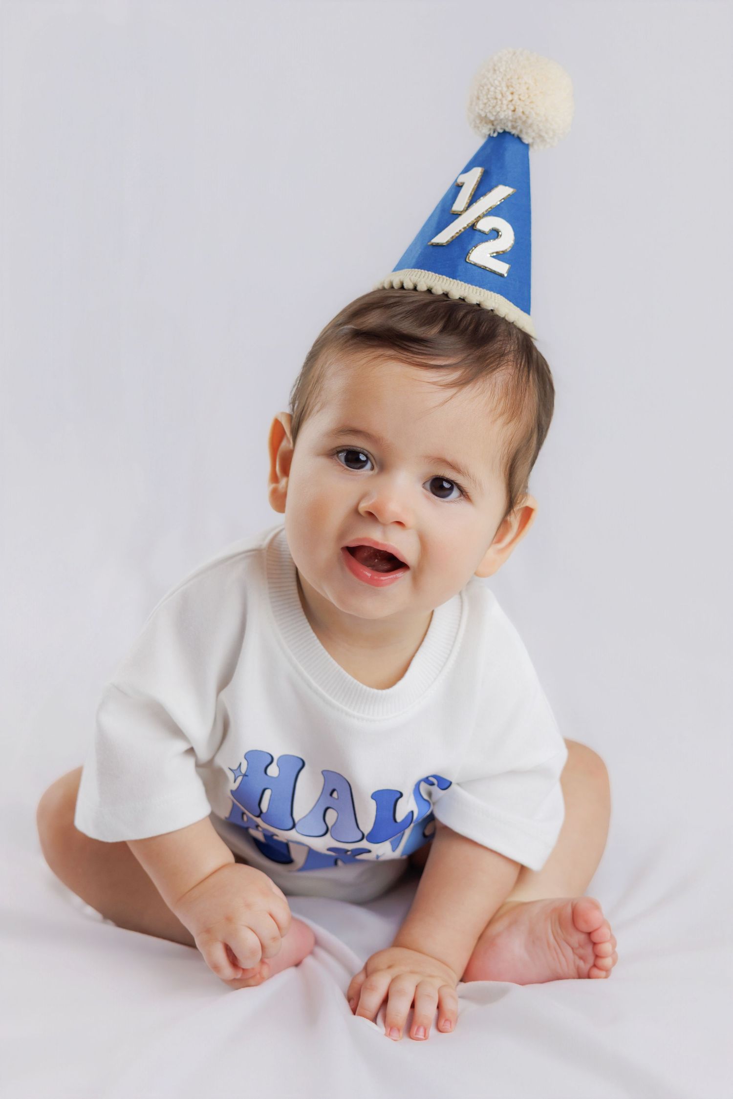 Adorable baby wearing a blue half birthday party hat and white onesie, sitting and smiling at camera.