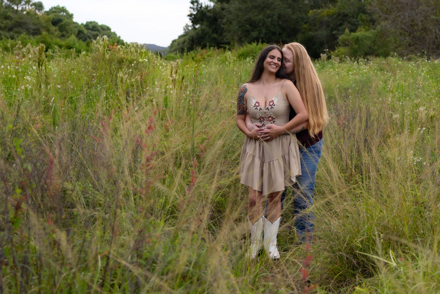 Two women smiling and embracing in a lush green meadow, one wearing a beige dress with white boots.
