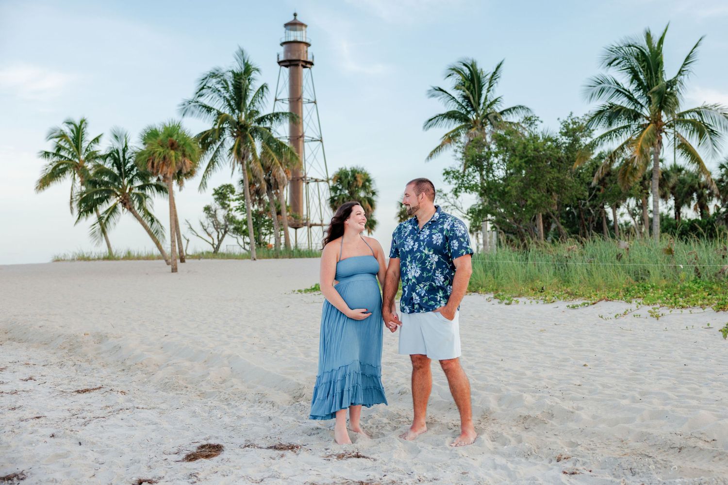 Expectant couple holding hands on a sandy beach maternity photo session with palm trees and a lighthouse in the background.