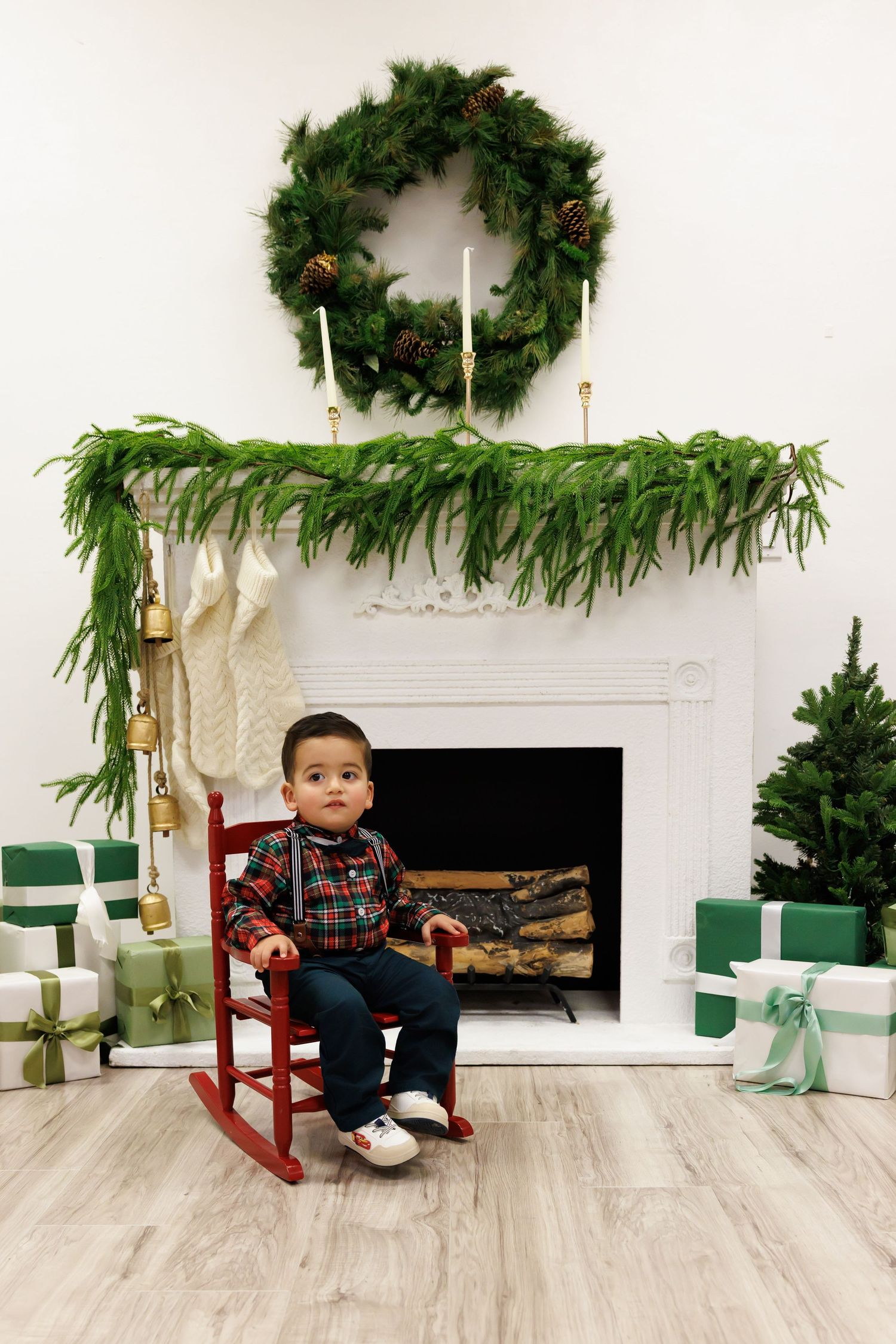 Toddler boy in plaid shirt sits in red rocking chair by white Christmas fireplace with wreath, garland, stockings and gifts.