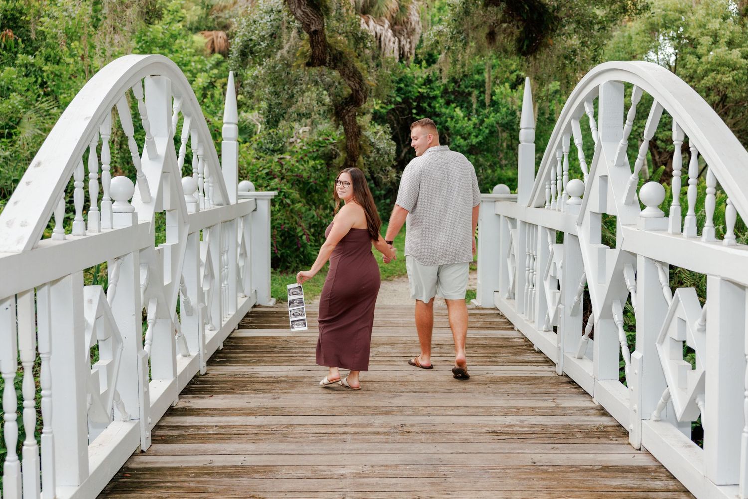 Couple holding hands walks across a white wooden footbridge surrounded by lush green trees while the woman looks back at the camera.