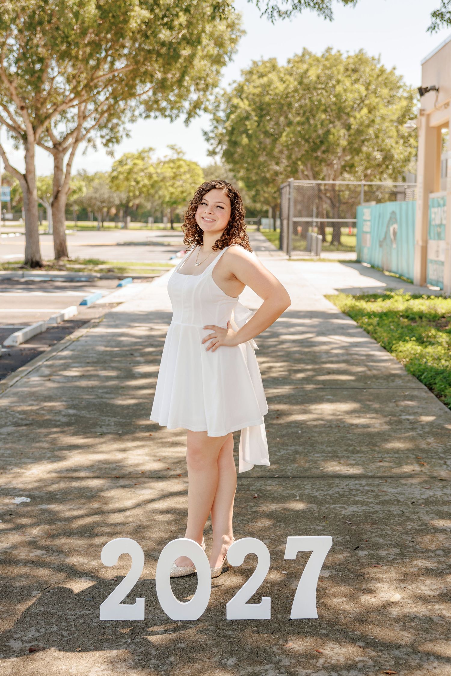 Smiling girl with curly hair in a white dress poses on a sunny sidewalk beside white 2027 numbers for her class of 2027 senior portrait.