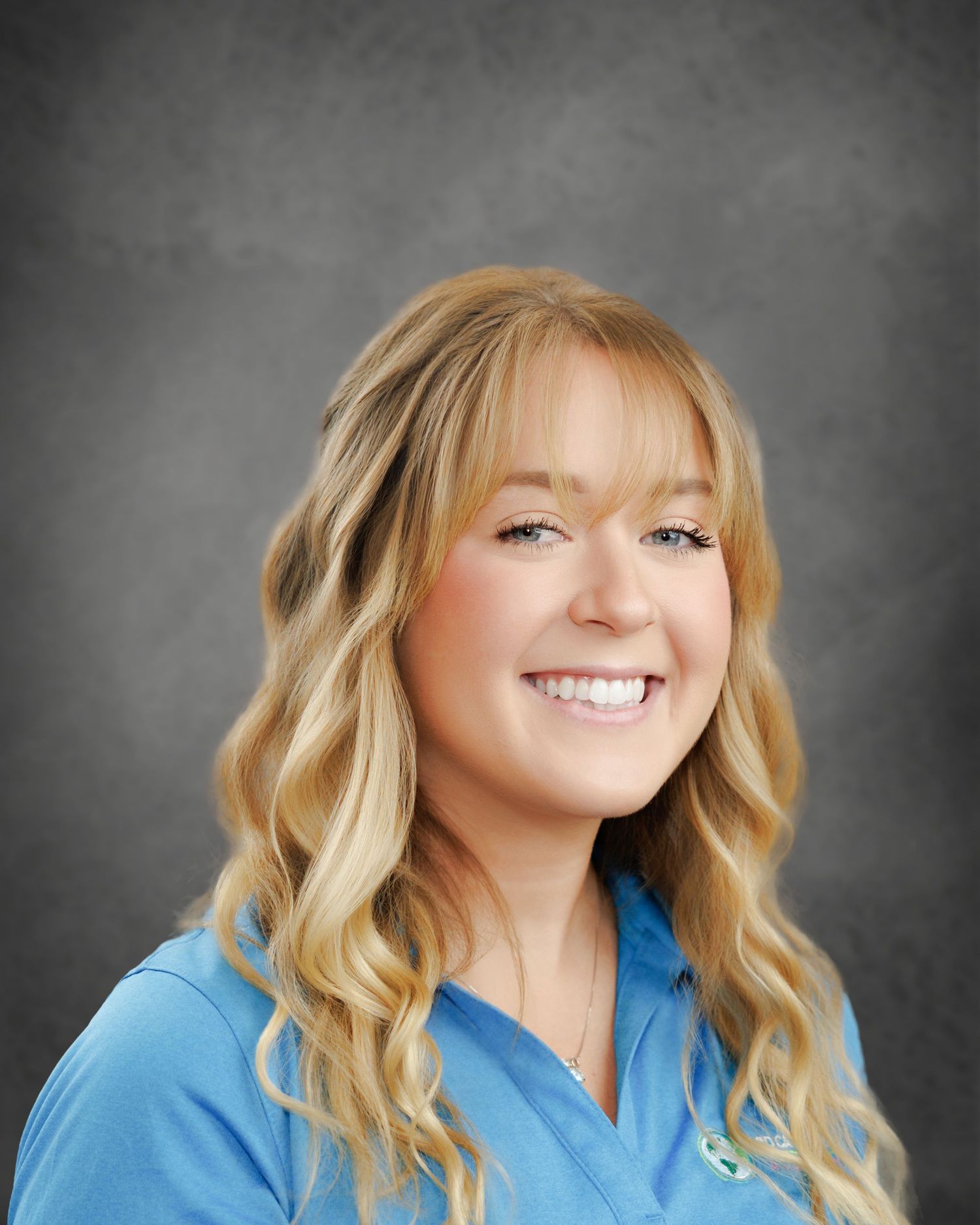 Smiling woman with long wavy blonde hair and bangs wearing a light blue polo shirt in a professional headshot against a gray background.