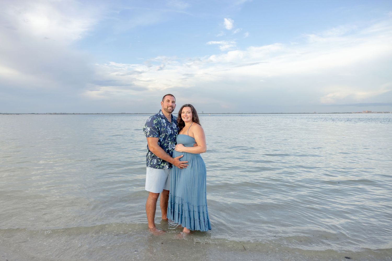 Couple in a blue dress and floral shirt stand barefoot in shallow beach water, embracing for a maternity photo under a soft cloudy sky.