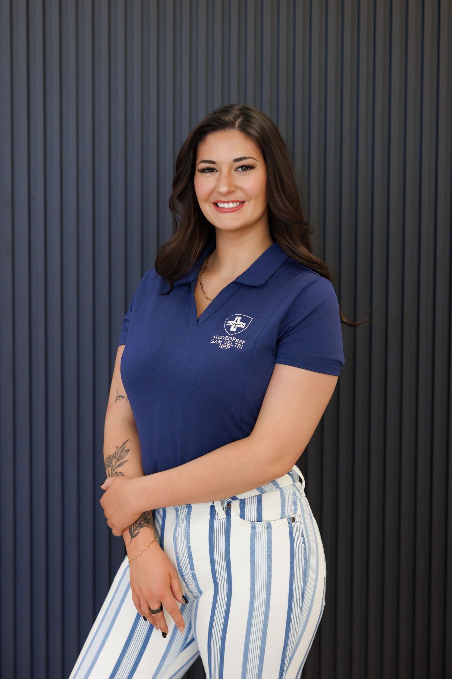 Smiling young woman in navy blue polo shirt and striped pants posing against dark corrugated metal wall.