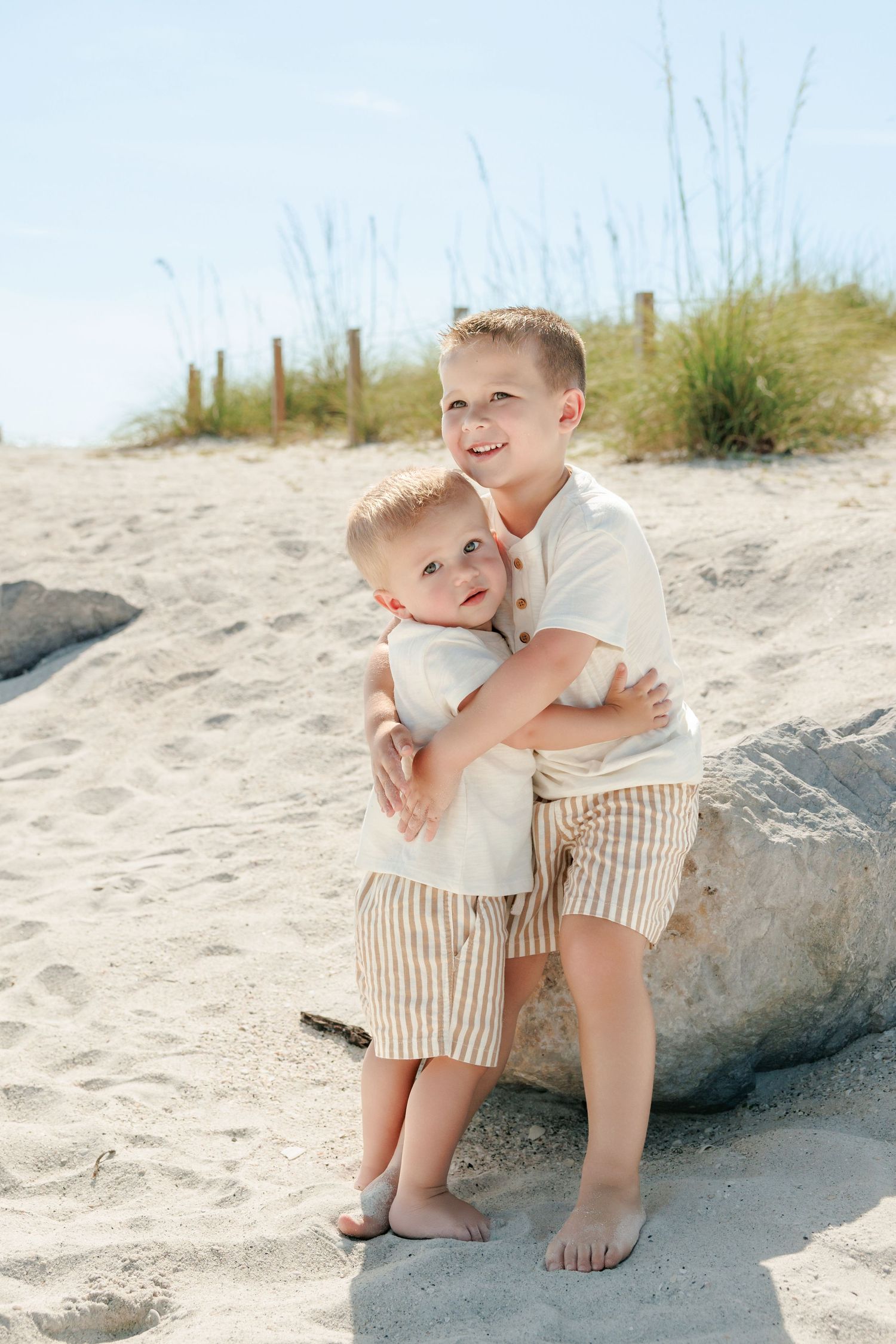 Two young brothers hugging on a sandy beach, wearing matching striped shorts and white shirts on a sunny day.