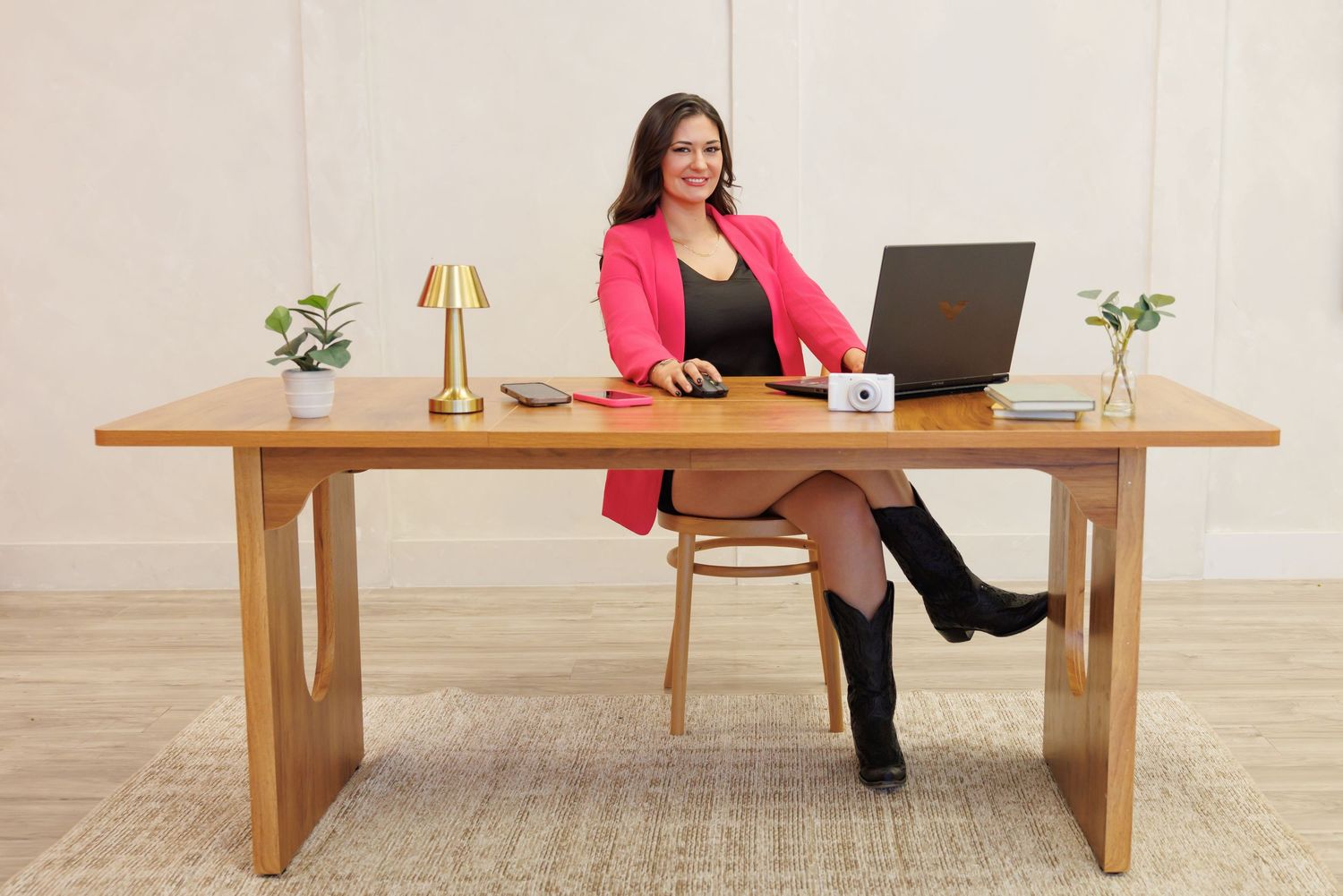 Woman in pink blazer sitting at wooden desk with laptop, lamp, and plants in bright office setting.