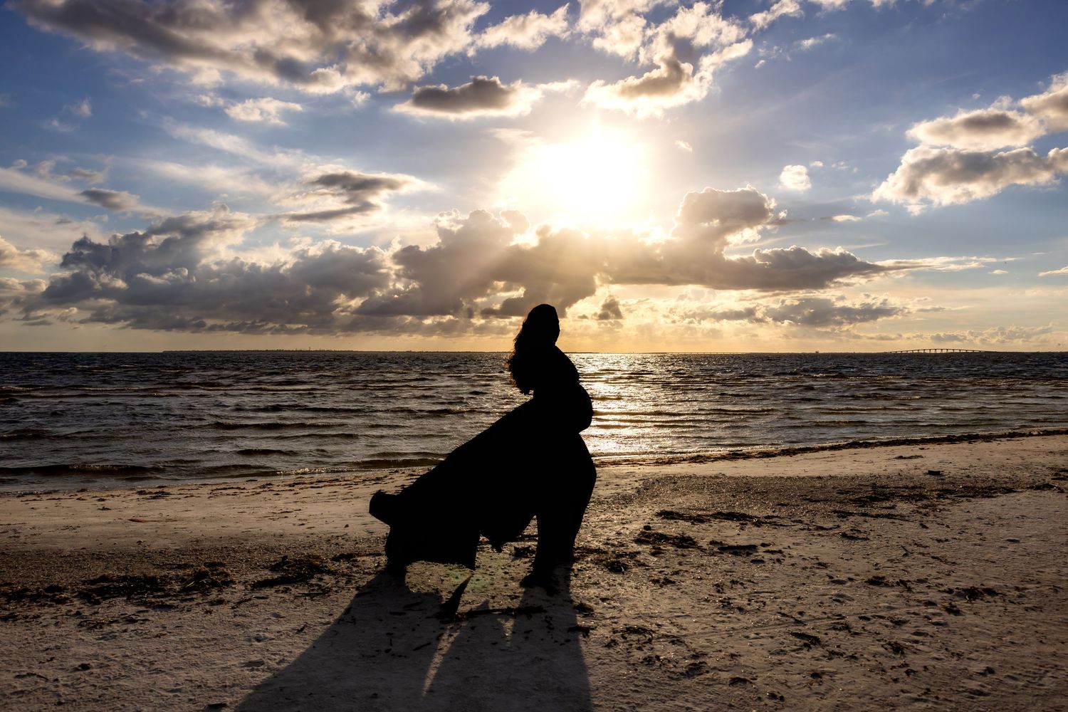 Silhouette of a woman in flowing dress walking on beach at golden sunset with dramatic cloudy sky.