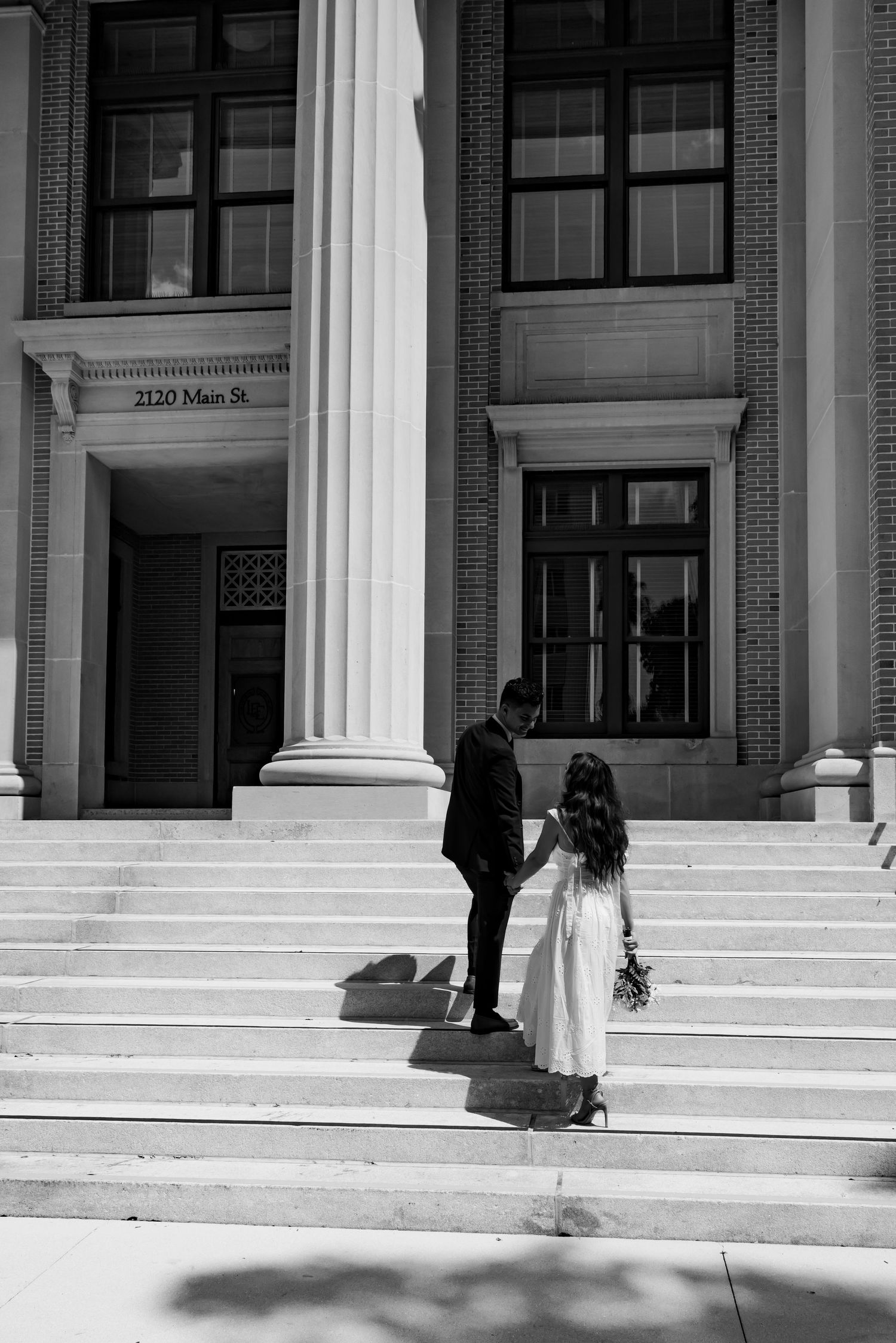 Black and white photo of a couple holding hands on steps of a brick building at 2120 Main St., bride holding bouquet.
