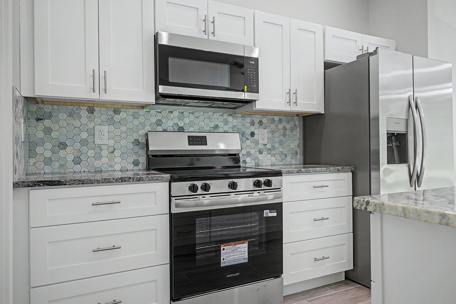 Kitchen with white shaker cabinets, gray granite countertops, mosaic tile backsplash, stainless steel range, microwave and refrigerator.