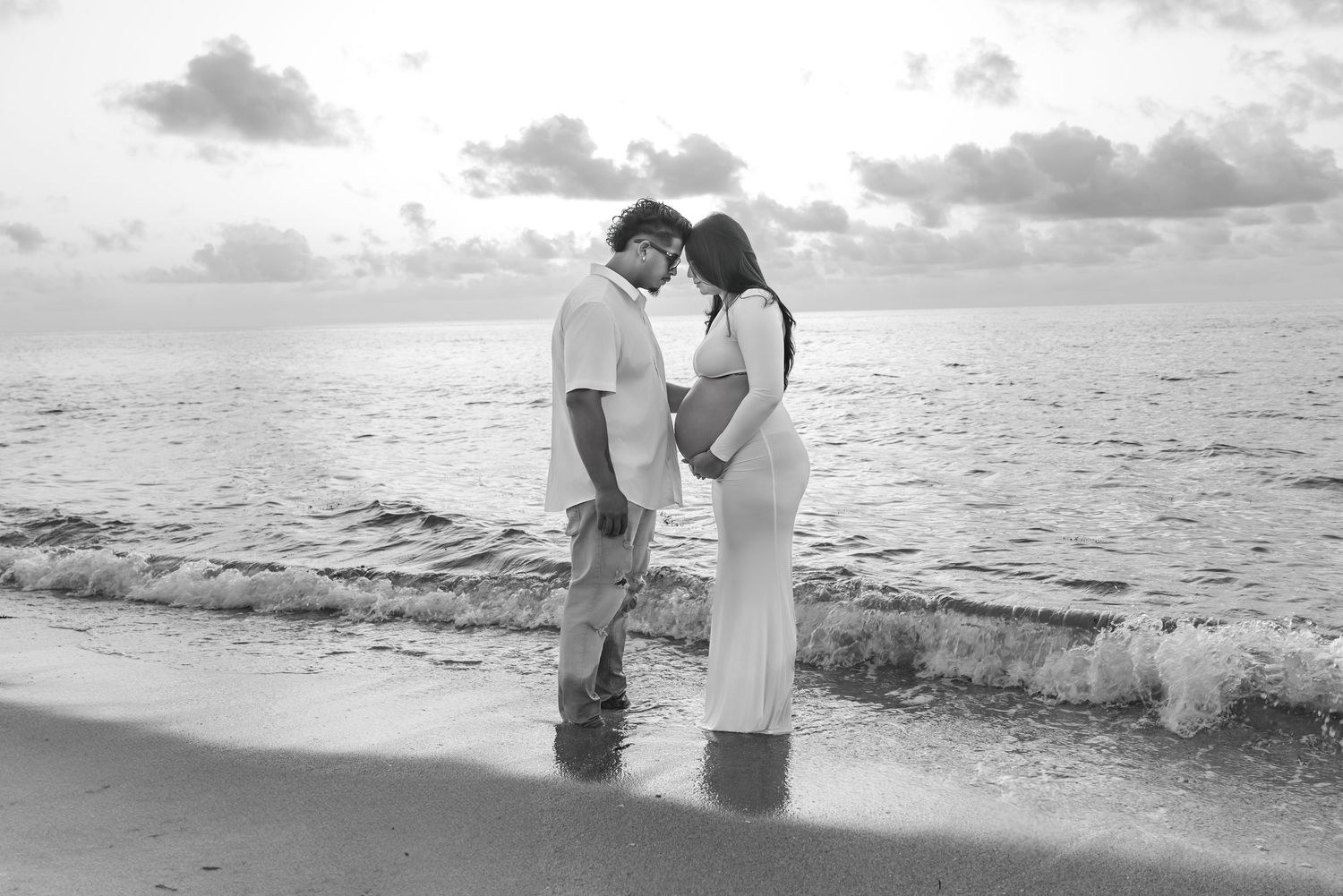 Black and white photo of a couple standing on a beach at sunset, facing each other near the ocean waves.