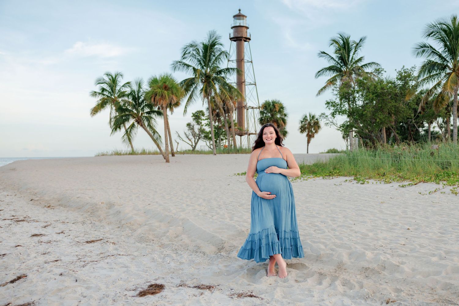 Pregnant woman in a flowing blue dress cradles her belly on a sandy beach with palm trees and a lighthouse in the background.