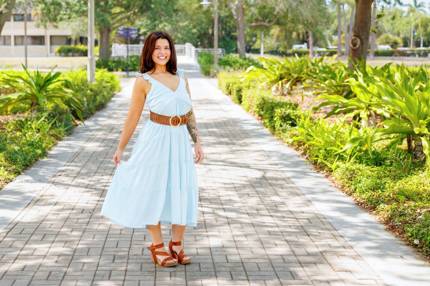 Smiling woman in a light blue sundress and brown belt walking along a brick paved path lined with tropical plants and palm trees.