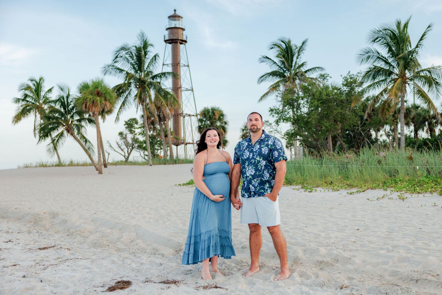 Expecting couple in a blue dress and floral shirt hold hands on a sandy beach with palm trees and a historic lighthouse behind them.